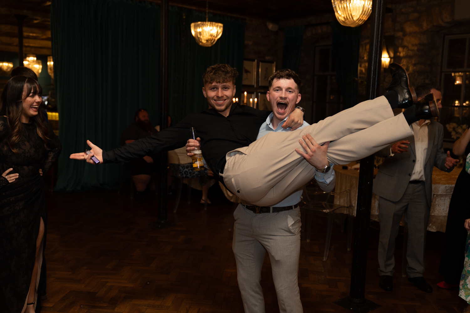 A groomsman carries his mate in his arms on the dance floor — both laughing, beer in hand, pure joy